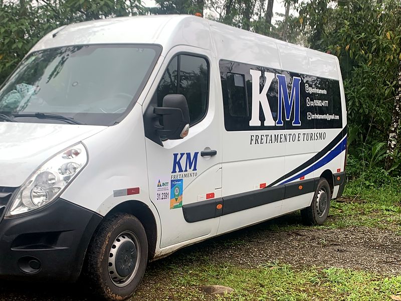 Van da KM Fretamento e Turismo vista de frente e de lado, em uma clareira de cascalho com mata ao fundo